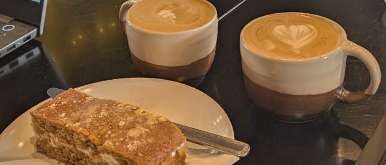 Two flat whites with latte art, a slice of cake and a laptop on a café table — Richard and Tiffany working together